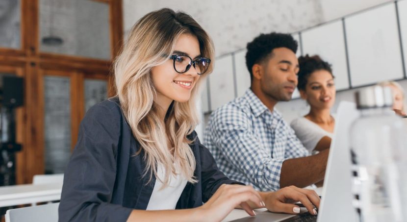 beautiful curly female freelancer with cute manicure using laptop smiling indoor portrait blonde secretary sitting beside african coworker blue shirt scaled 1