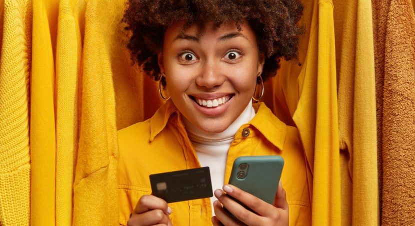 positive mixed race shopaholic stands near rack with clothes uses credit card smartphone making purchasing